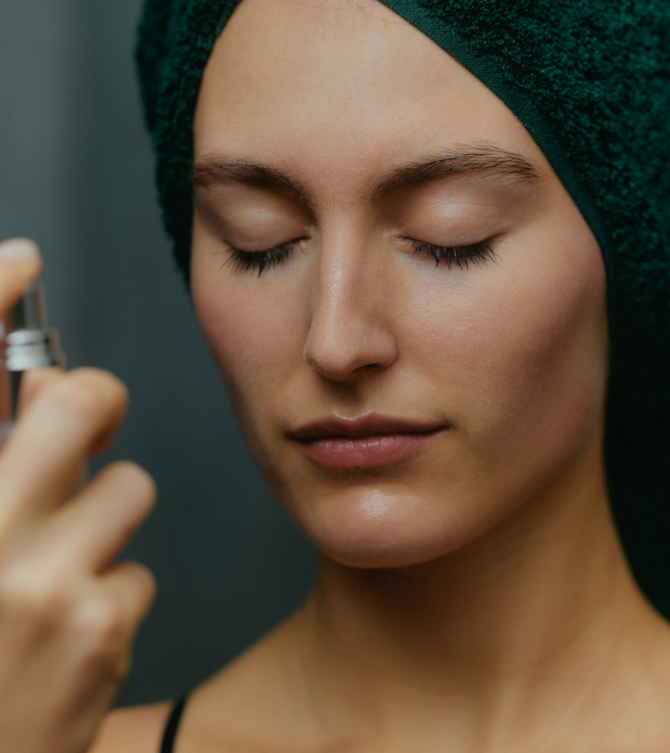 Woman with clean, makeup-free skin and a towel on her head, applying a facial mist with closed eyes. Hydrating and refreshing skincare routine for healthy, glowing skin.