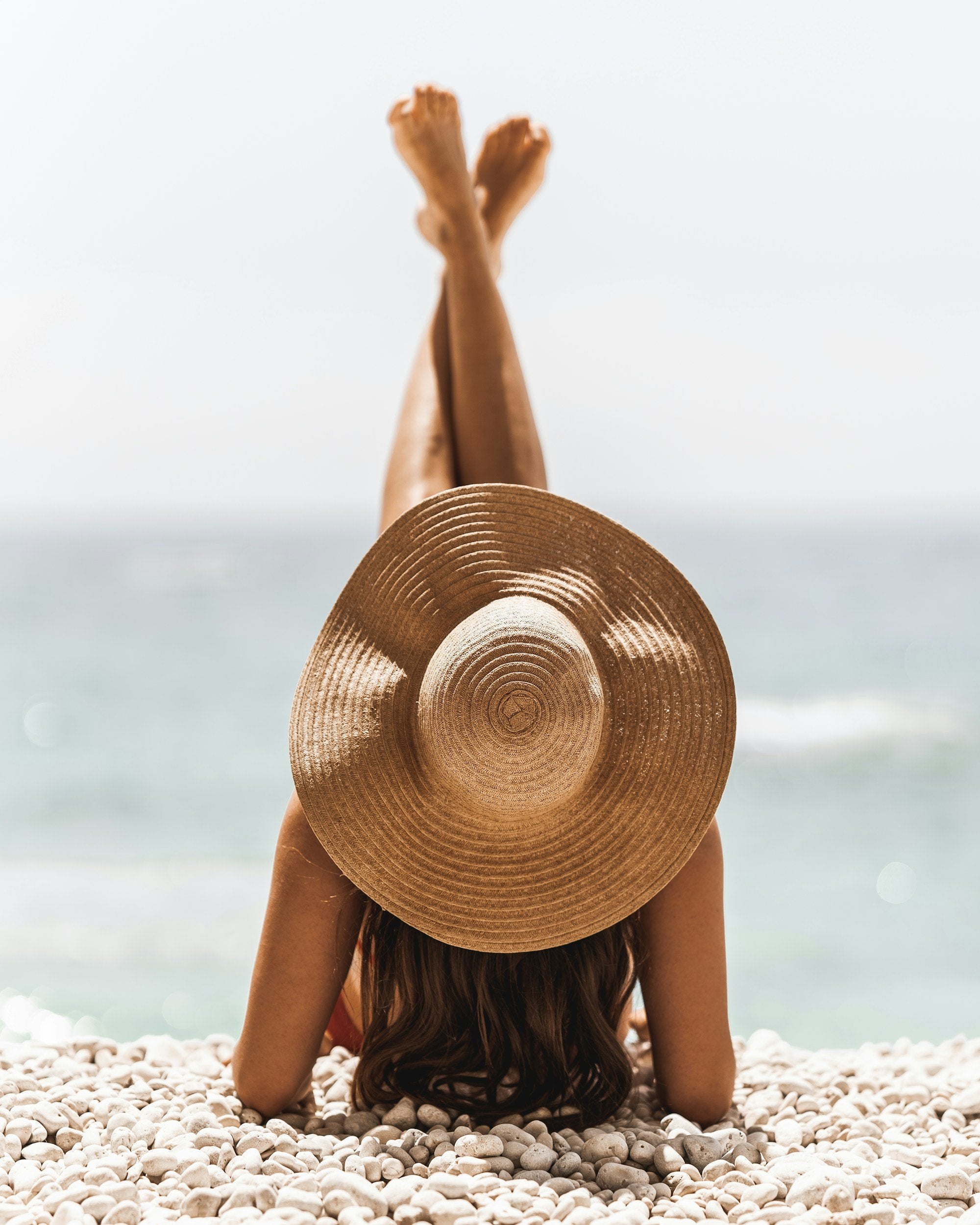 A woman sunbathing at the beach while wearing a large straw hat, protecting her face from UV rays. This image highlights the importance of sun protection to prevent skin damage, premature aging, and skin cancer.