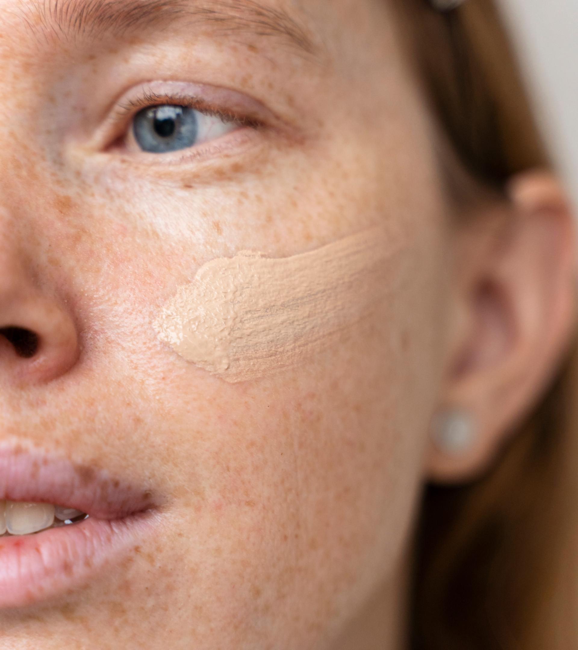 Close-up image of a young woman with red hair, freckles, and blue eyes, showing a swatch of foundation on her cheek. The image highlights her natural skin tone and the makeup product for a dermatology or beauty-related context.